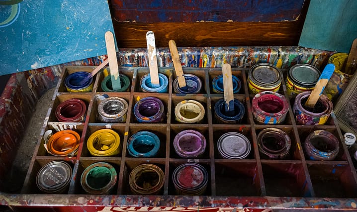 Top view of opened cans of dried up used colorful paints a few with stir sticks and some still closed grouped together in a wooden box with dividers