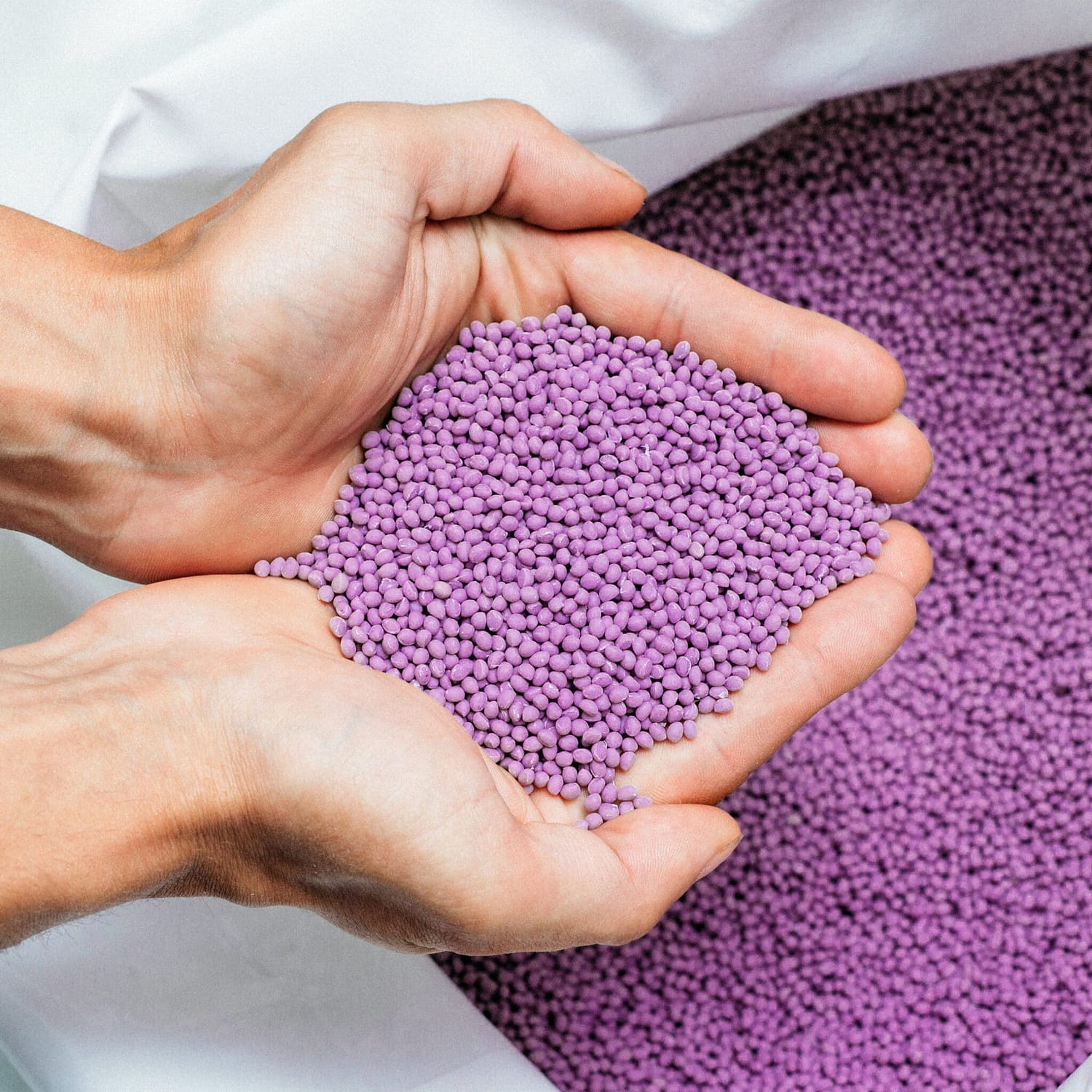A bag of purple plastic pellets sits in the background. Hands scoop out a handful of pellets to bring them in screen.