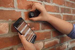 The Nix Mini is held up to scan a brick wall. A phone is also being held, showing the scan results.